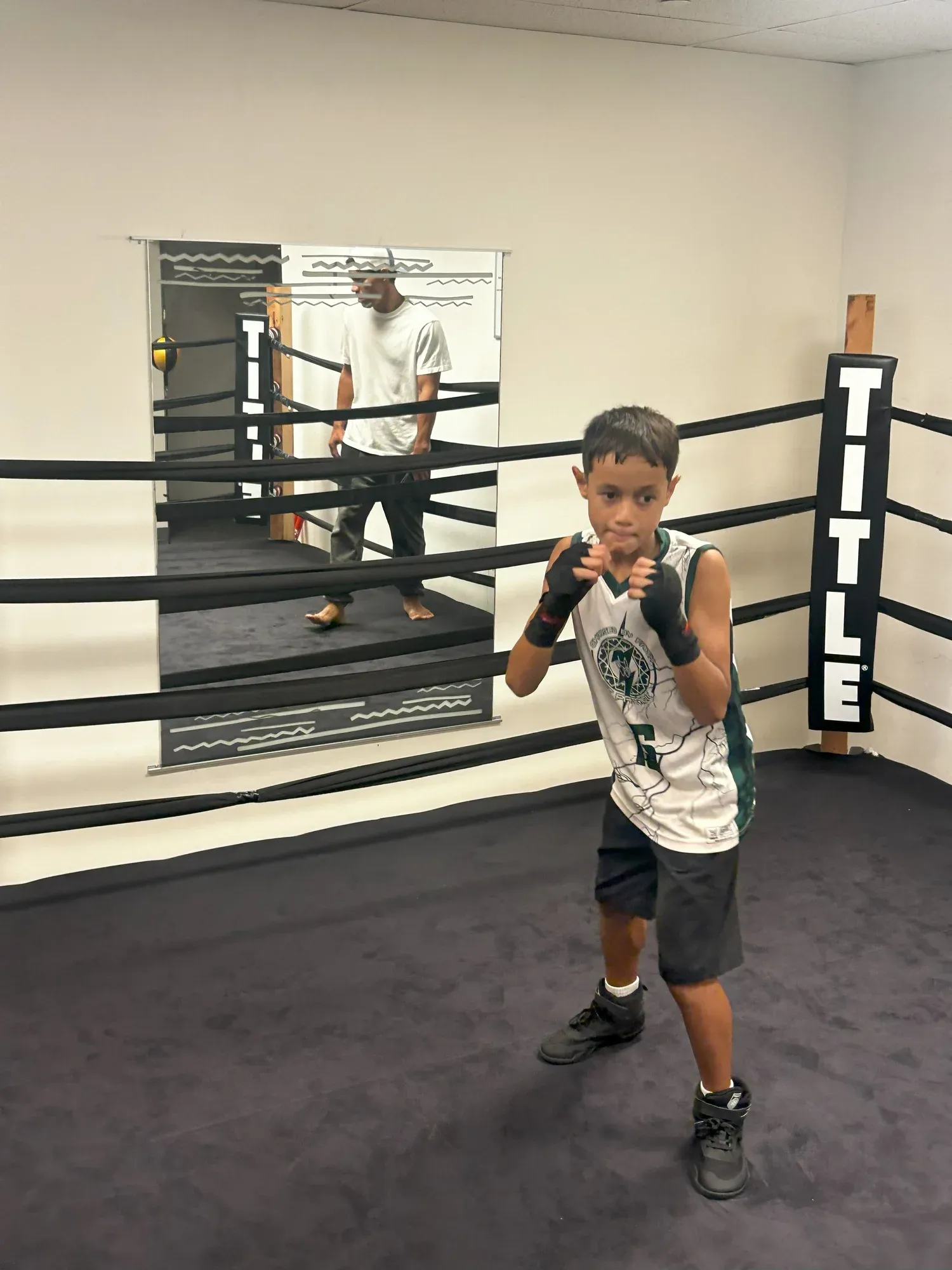 A young boxer in fighting stance inside the Iron Kingz ring, coach reflected in the mirror